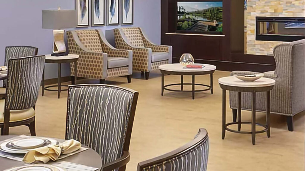 A modern, well-lit lounge area with patterned chairs, round tables, a television, framed art on the wall, and a decorative lamp, creating a comfortable and inviting atmosphere.