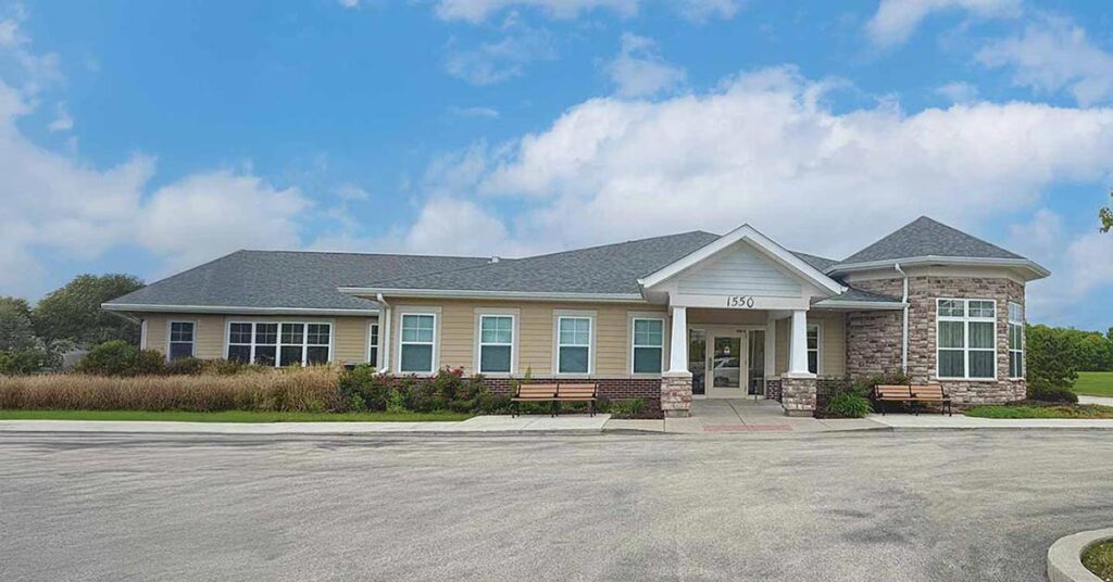 One-story building with light-colored siding, stone accents, and a gray roof, labeled 1350. Benches are placed near the entrance, and landscaped bushes line the front. Blue sky with scattered clouds is above.