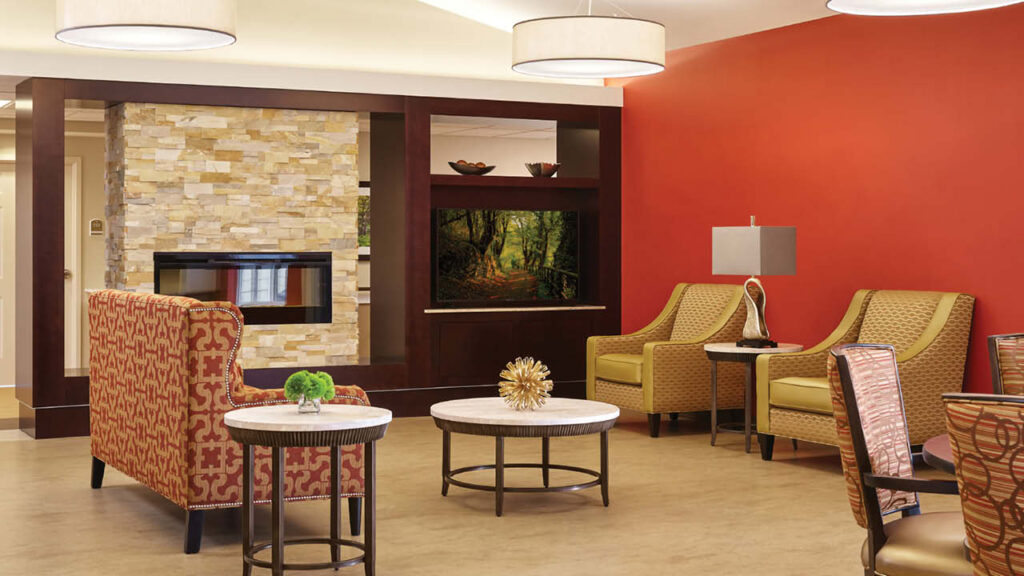 A modern lounge area with patterned armchairs, marble-top tables, a stone fireplace, a flat-screen TV, and a vibrant red accent wall, decorated with lamps and small green plants.