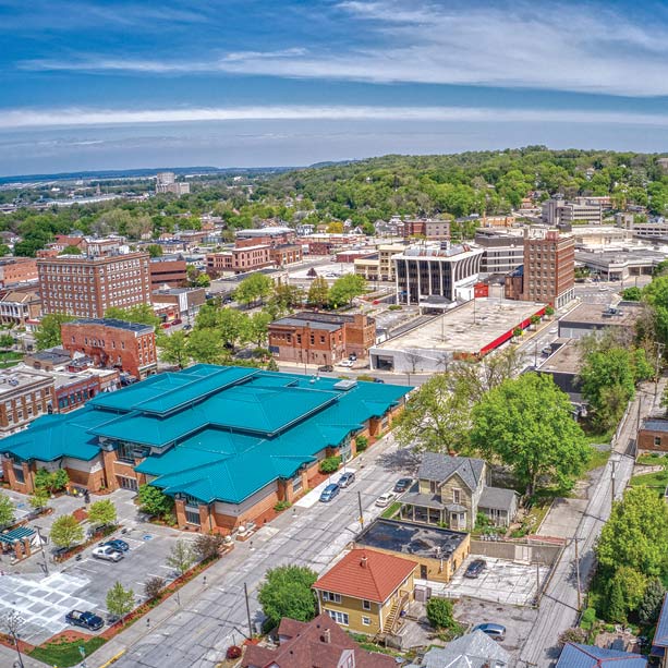 Aerial view of a small city with a mix of commercial buildings, houses, and tree-lined streets under a partly cloudy sky. Green roofs and a hilly, forested area are visible in the background.