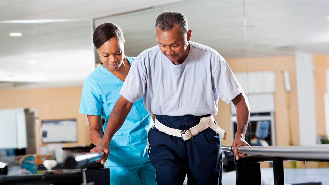 A healthcare professional assists a man in physical therapy as he walks using parallel bars. The man wears a support belt, and the setting appears to be a rehabilitation facility.