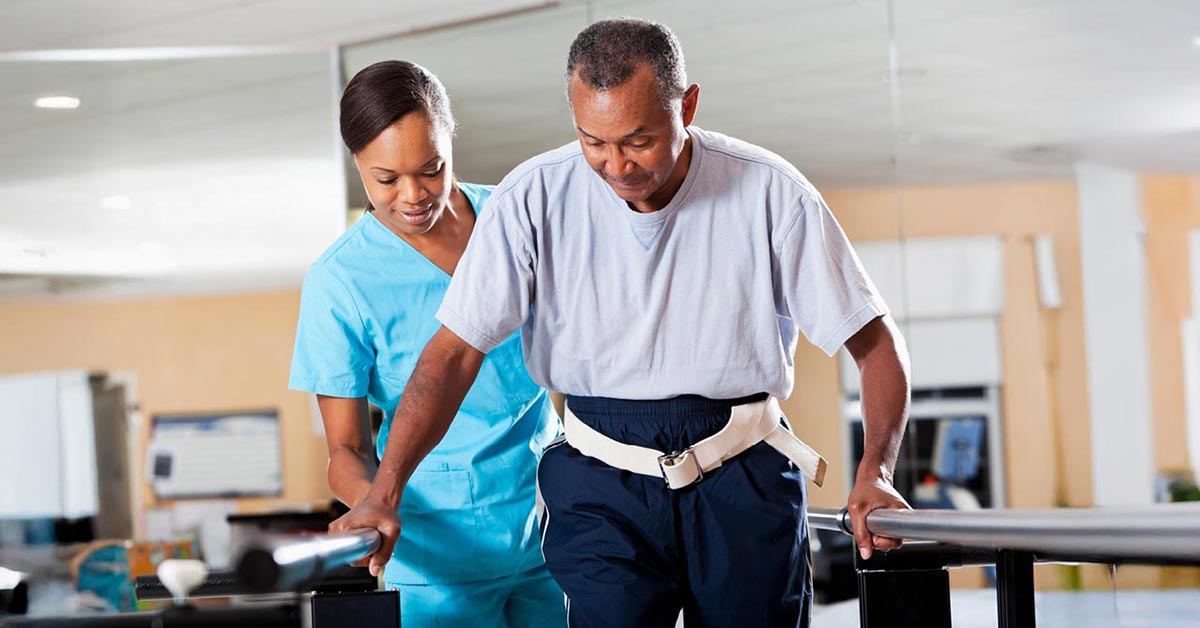 A nurse in blue scrubs assists an older man in a gray shirt and gait belt as he practices walking using parallel bars in a rehabilitation facility.