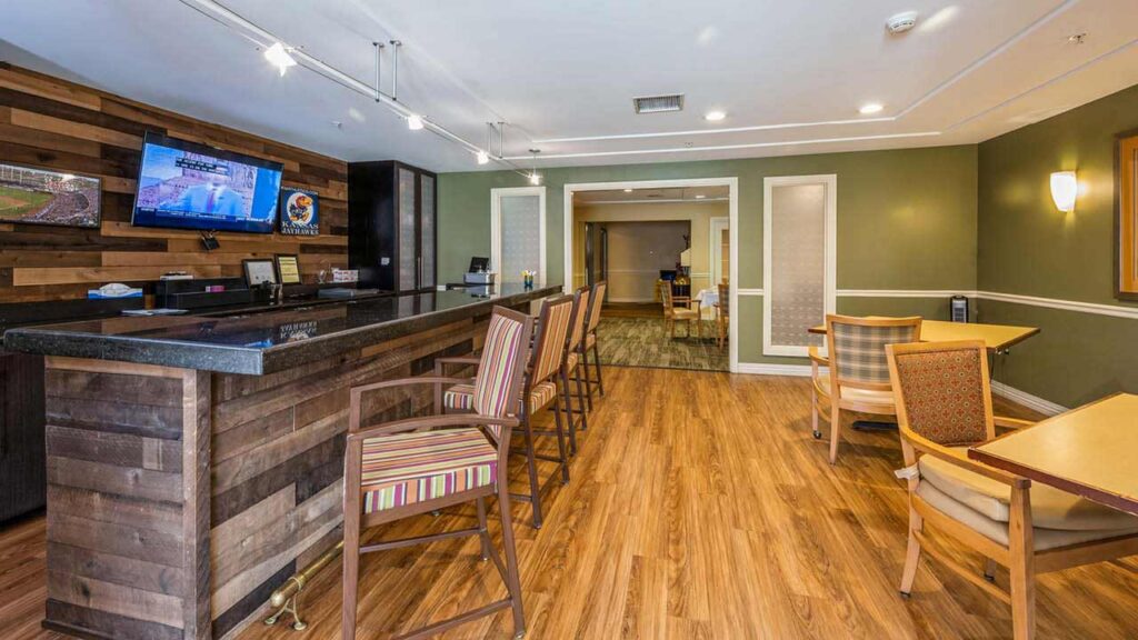 A cozy lounge area with a wooden bar counter, striped bar stools, and tables with cushioned chairs on a wood floor. A TV is mounted on the wall, and the walls are painted green and brown.