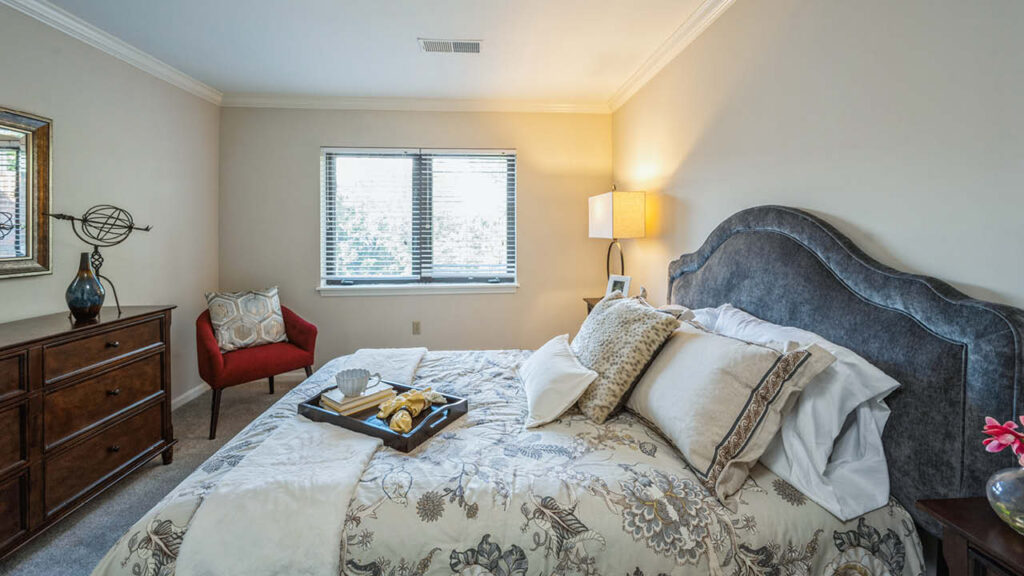 A cozy bedroom with a large bed featuring patterned bedding and multiple pillows, a tray with breakfast items, a wooden dresser, a red accent chair, a lamp, and a window allowing in natural light.