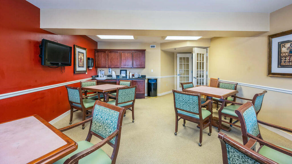 A small dining or activity room with four square tables and green cushioned chairs, a wall-mounted TV, cabinets, a counter with appliances, and red and beige walls. Framed artwork decorates the walls.