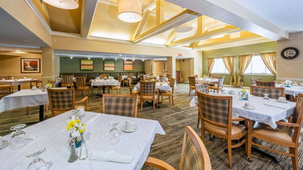 A bright, spacious dining room with neatly set tables, wooden chairs, large windows with curtains, and soft lighting. The walls are decorated with framed art, and yellow flowers are on some tables.
