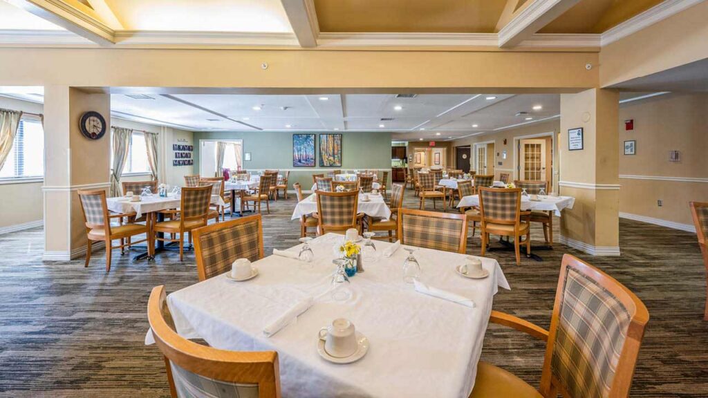 A spacious, well-lit dining room with several wooden tables and chairs, set with white tablecloths, napkins, cups, and glasses. Large windows and wall art add a bright, welcoming atmosphere.