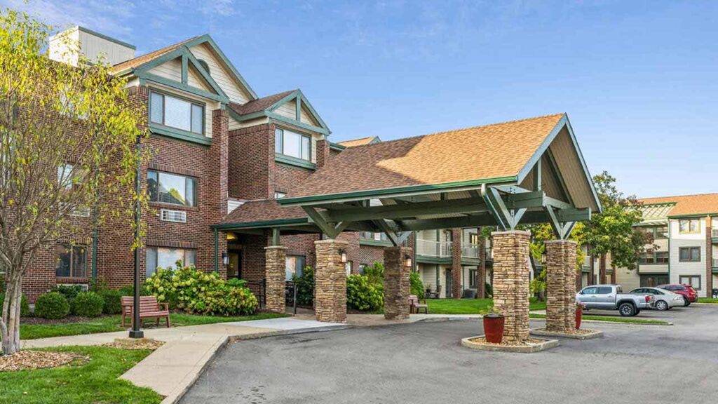 A brick building with large windows and a covered entrance supported by stone pillars, surrounded by greenery, with a parking lot and parked cars in front under a blue sky.