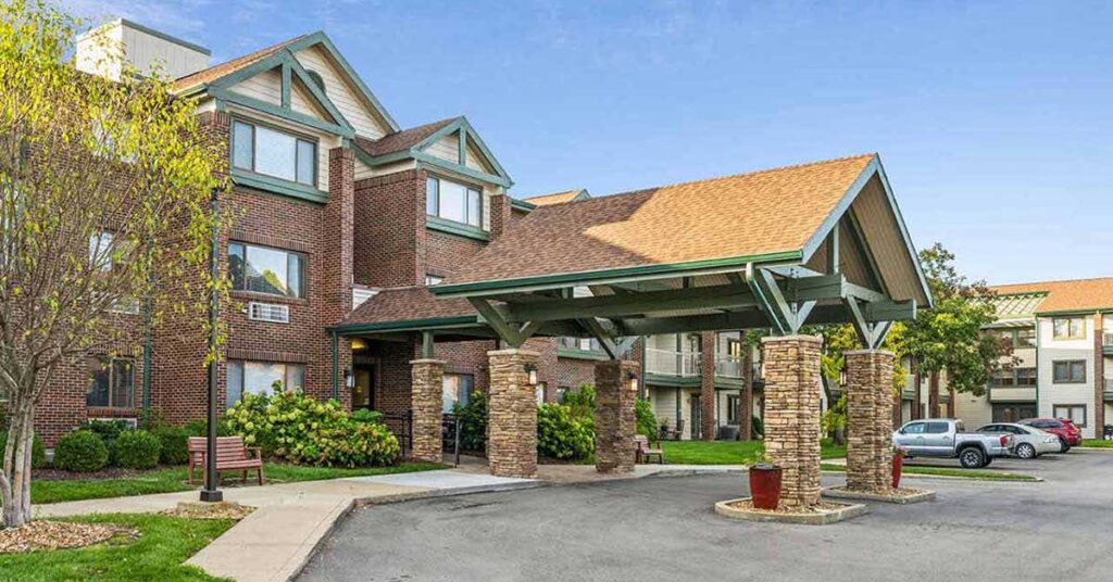 A brick building with a green-roofed entrance canopy supported by stone columns, surrounded by landscaping, trees, and parked cars under a clear blue sky.