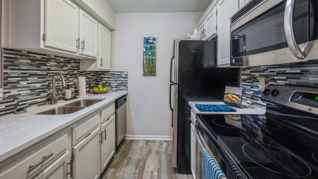 Modern kitchen with white cabinets, stainless steel appliances, black refrigerator, electric stove, double sink, and gray-blue mosaic tile backsplash. Fruit bowl and cookbook sit on the white countertop.