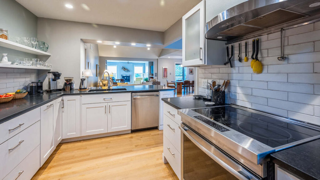 Modern kitchen with white cabinets, black countertops, stainless steel appliances, open shelves, and a tile backsplash. The space opens to a living and dining area with wood flooring and bright, natural light.