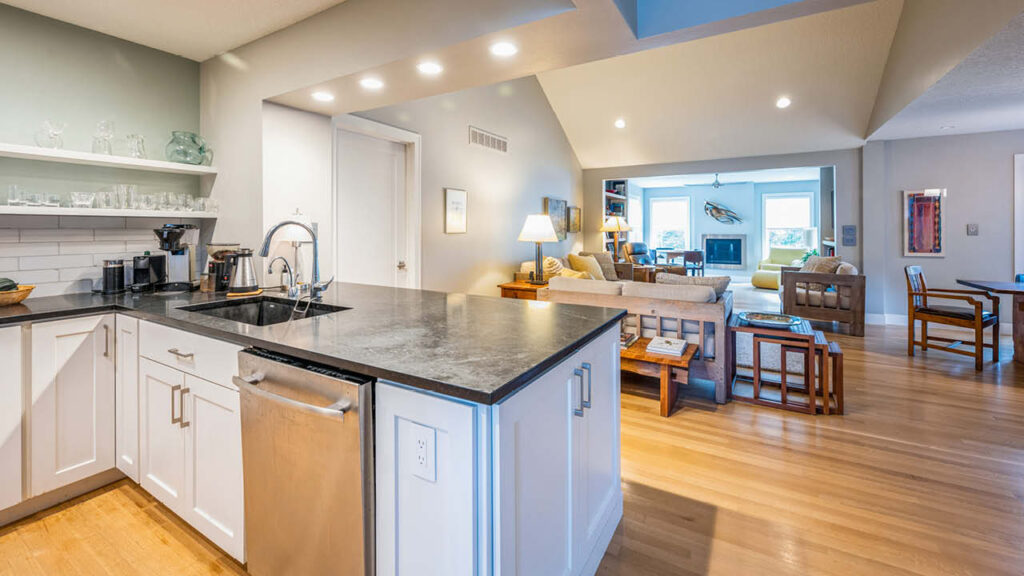 Open-concept kitchen with white cabinets, black countertops, and stainless steel appliances, overlooking a bright living room with wooden floors, modern furniture, and large windows letting in natural light.