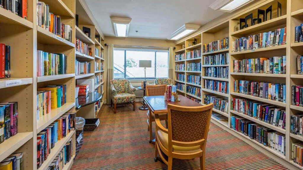 A cozy library room with bookshelves on both sides, a wooden table and two chairs in the center, and two armchairs by a large window letting in natural light.
