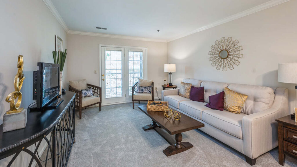 A bright living room with a beige sofa, purple and gold pillows, two patterned armchairs, a wooden coffee table, and a TV on a console. French doors let in natural light. Decor includes a sunburst mirror and modern accents.