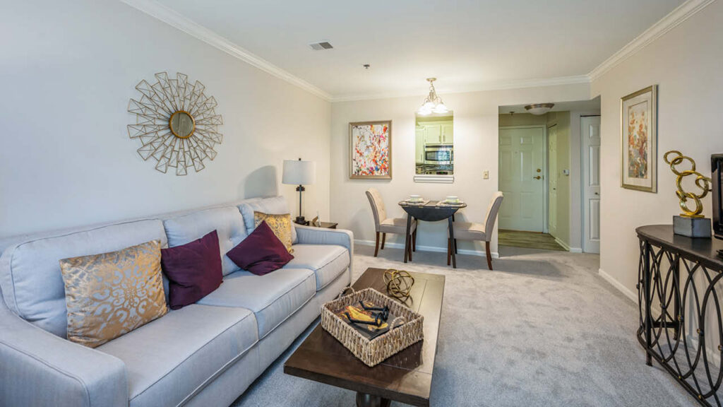 A cozy living room with a light gray sofa, gold and purple pillows, a wooden coffee table, and modern decor. In the background, a small dining table for two is set near the kitchen entrance.