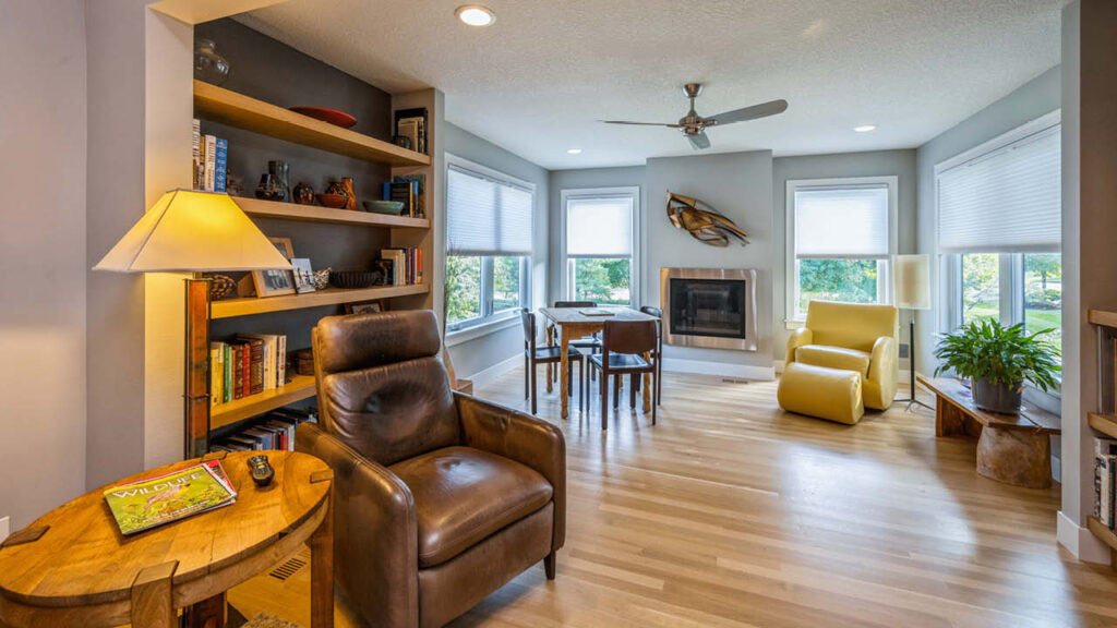 A cozy living room with wooden floors, a leather armchair, bookshelf, round table with four chairs, yellow armchair, fireplace, ceiling fan, and large windows letting in natural light.