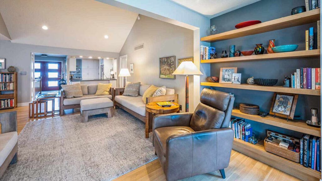 A cozy living room with a gray armchair, beige sofas, a large area rug, wooden bookshelves filled with books and decor, and warm lighting, leading to a view of the kitchen and entryway in the background.