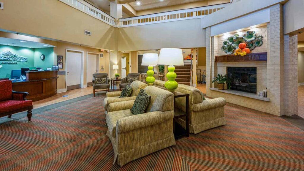 Hotel lobby with striped carpet, beige sofas, green lamps, a fireplace decorated with plants and pumpkins, wooden reception desk, and chairs. The area is well-lit and spacious with a second-floor balcony visible.