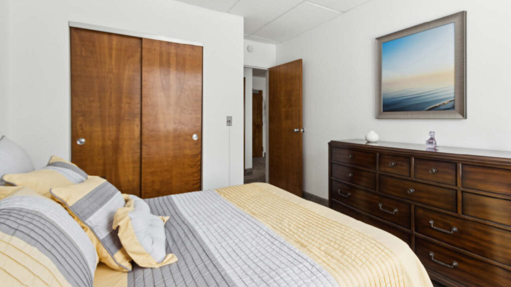 A neatly made bed with gray and yellow bedding sits in a bedroom with wood sliding closet doors, a wooden dresser, and a framed seascape picture on a white wall.