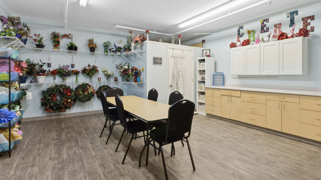 A bright craft room with a wooden table and five black chairs, shelves of artificial flowers and wreaths, white cabinets, and the word 