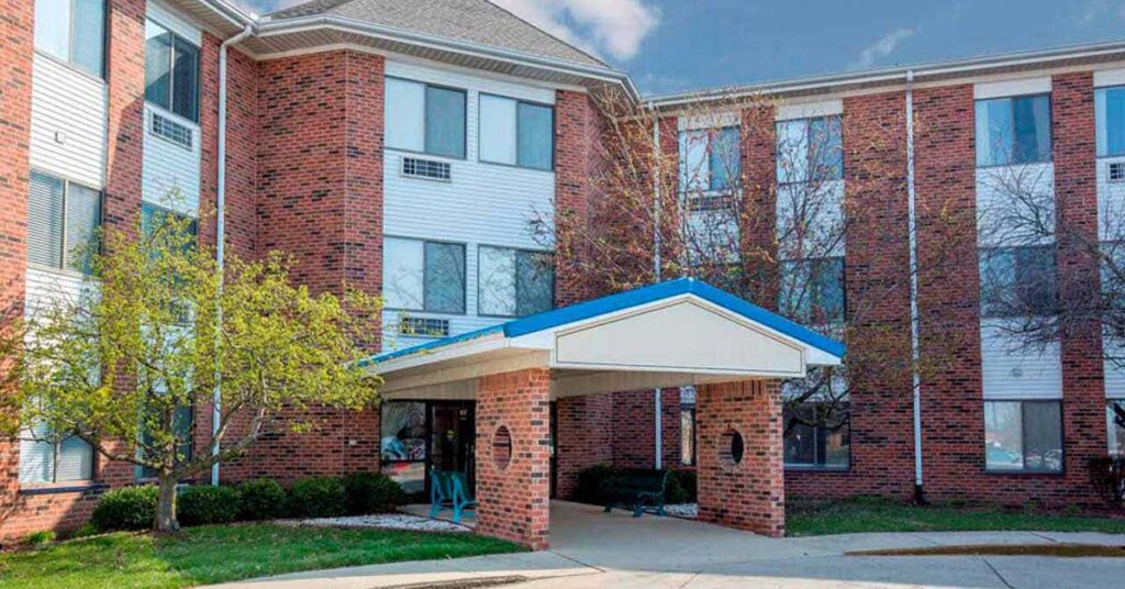 A three-story brick building with white siding, featuring a covered entrance supported by brick columns. There are trees and shrubs near the entrance, and a clear, blue sky in the background.