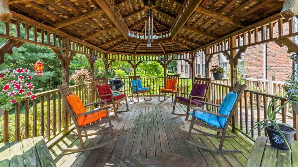 A wooden gazebo with a vaulted roof, featuring multiple rocking chairs with colorful cushions arranged in a circle. Bright flowers and greenery are visible outside the railing on a sunny day.