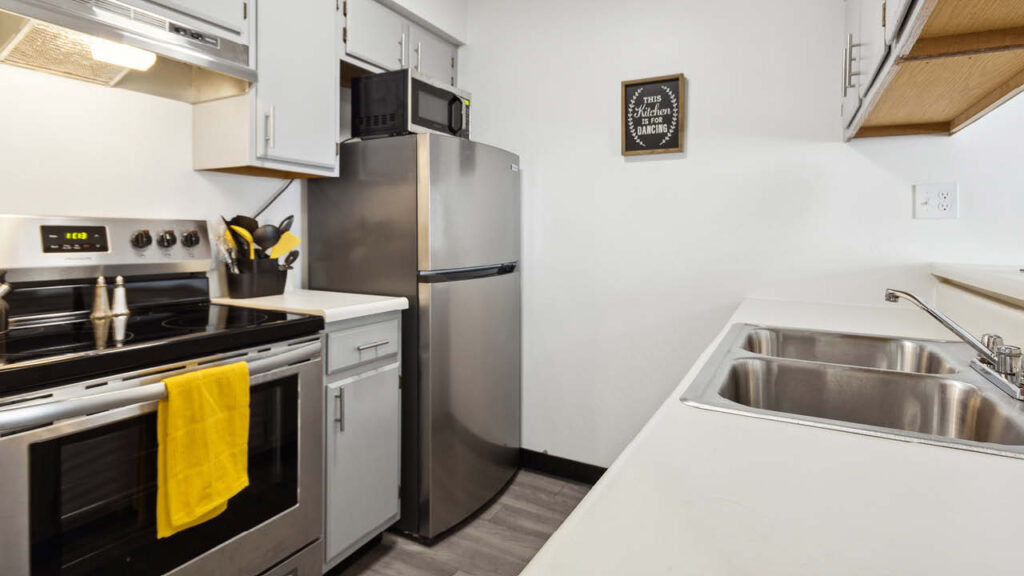 A modern kitchen with stainless steel appliances, white cabinets, a double sink, a yellow towel hanging on the oven, and utensils by the stove. A framed quote hangs above a microwave on top of the refrigerator.