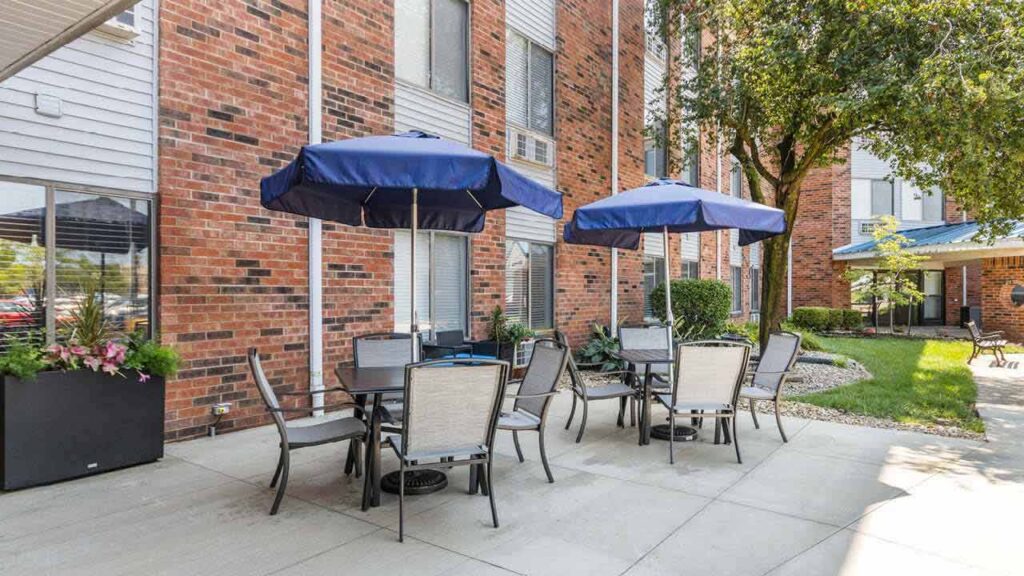 Outdoor patio with several tables and chairs under blue umbrellas, next to a red brick building. There are some potted plants and trees, with a grassy area and pathway in the background.