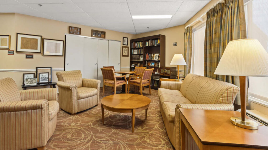 A cozy lounge with striped armchairs, a sofa, a round coffee table, a wooden dining table with chairs, bookshelves, framed pictures on the wall, and two table lamps, all in warm, neutral colors.