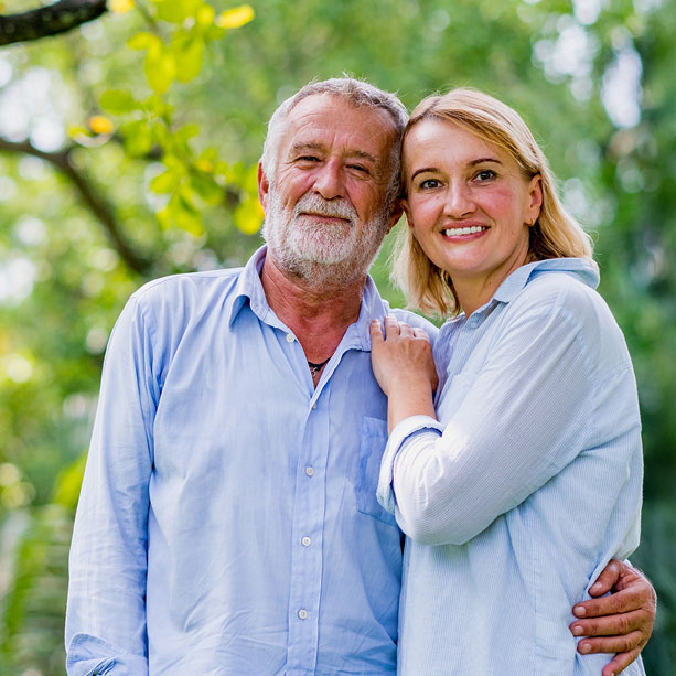 An older man and a younger woman stand close together outdoors, smiling at the camera. They are both wearing light blue shirts, and greenery is visible in the blurred background.