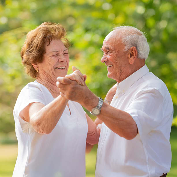 An elderly couple wearing white shirts smiles and dances together outdoors, surrounded by greenery and sunlight.