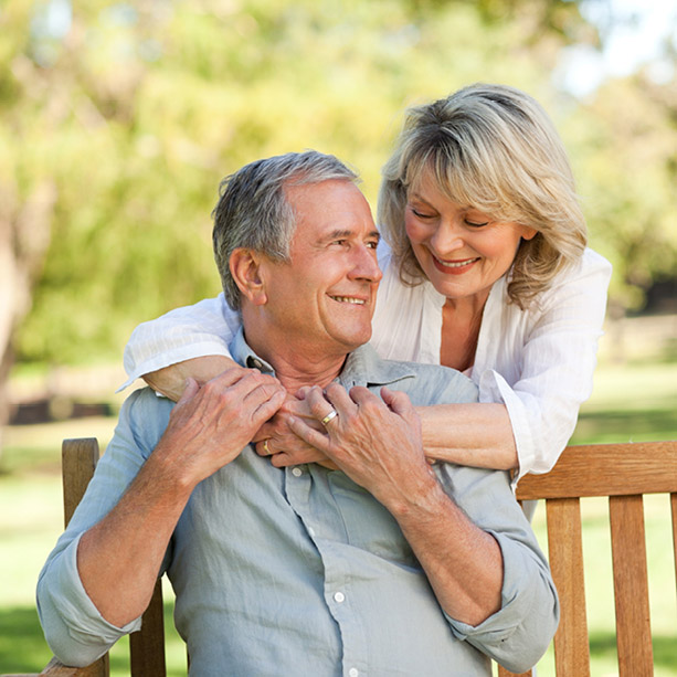 An older couple sits on a wooden bench outdoors, smiling at each other. The woman stands behind the man, embracing him with her arms around his shoulders. They appear happy and relaxed in a sunny park setting.
