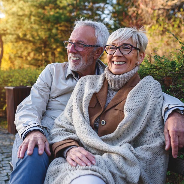 An older man and woman sit close together on an outdoor bench, smiling and laughing. The woman is wrapped in a knit blanket, and both wear glasses. Green trees and sunlight are visible in the background.