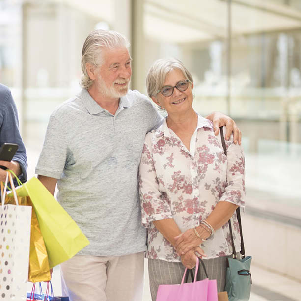 An older couple stands close together, smiling and holding shopping bags. The man has his arm around the woman. They are indoors, possibly in a mall, and appear happy and relaxed.