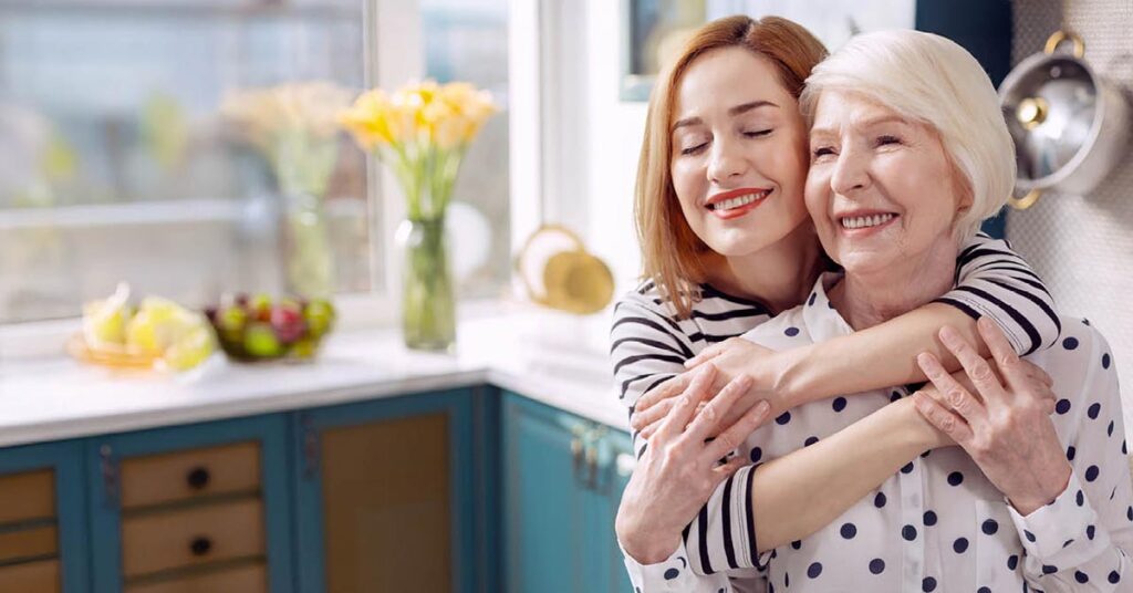 A young woman hugs an older woman from behind in a bright kitchen. Both are smiling and appear happy, with sunlight streaming in and flowers on the counter in the background.