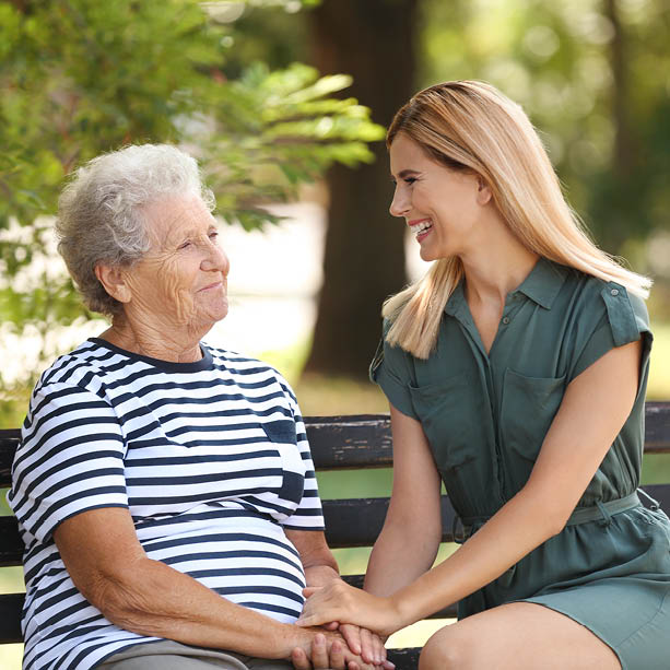A smiling young woman in a green dress sits on a bench holding hands and laughing with an older woman in a striped shirt, outdoors on a sunny day.