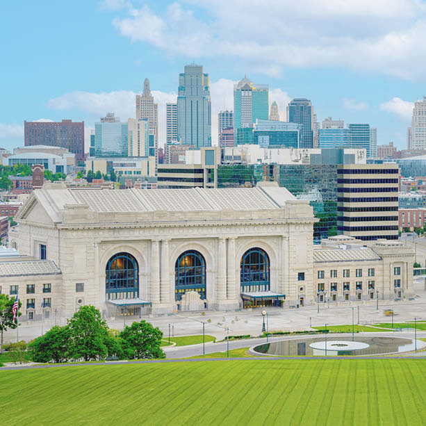 Union Station in Kansas City stands in the foreground with its grand arches, while modern skyscrapers and city buildings rise in the background under a partly cloudy blue sky.