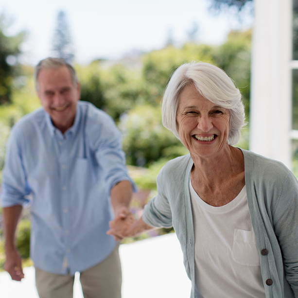 An older woman smiles brightly while holding hands with an older man outdoors. Both are casually dressed and appear happy, with greenery and sunlight in the background.