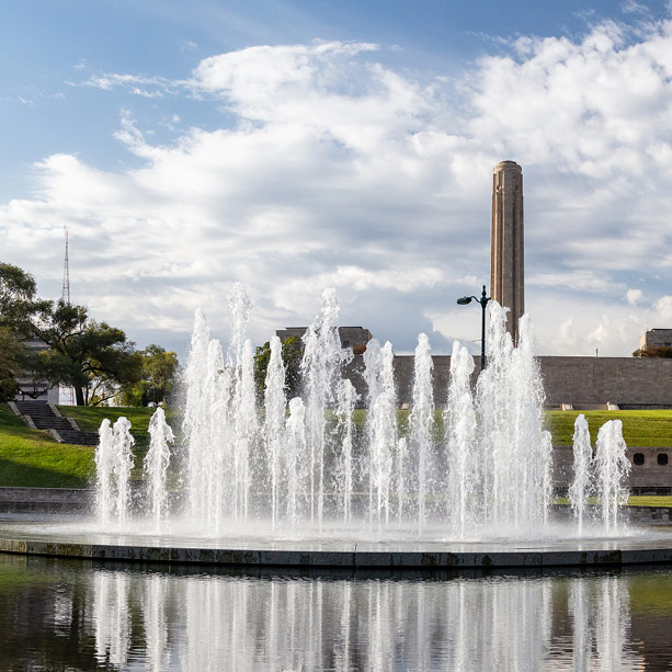 A circular fountain surrounded by water sprays upward in a park, with a tall stone tower and grassy areas under a partly cloudy sky in the background.