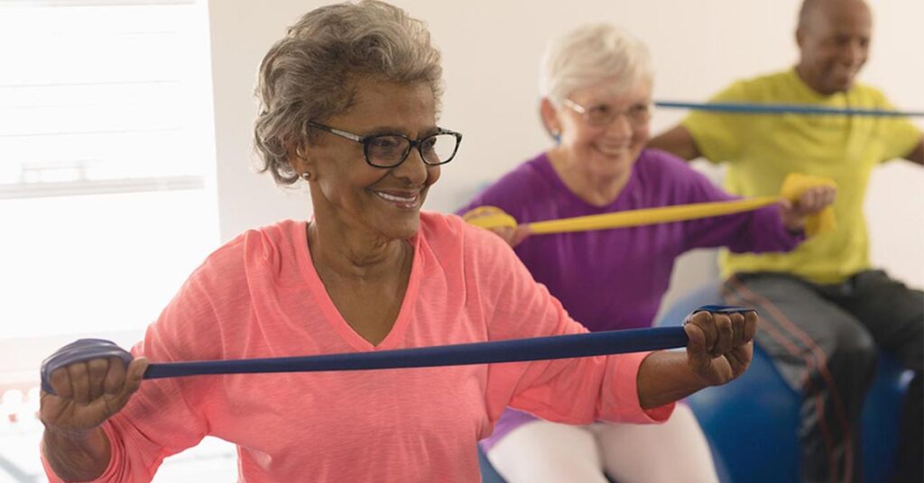 Three older adults exercise indoors, smiling as they use resistance bands. The woman in front wears glasses and a pink top, while the others behind her wear yellow and white tops. They appear engaged and happy.