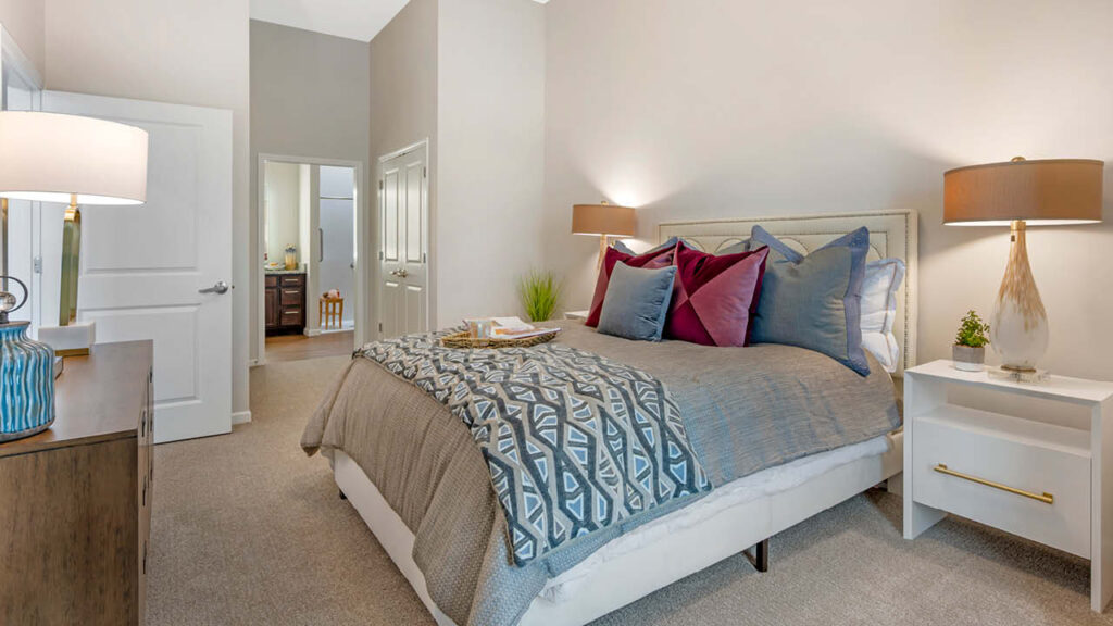 Modern bedroom with a large bed featuring patterned bedding and colorful pillows, flanked by white nightstands with lamps. A dresser and lamp are on the left, and an open door leads to a hallway and another room.
