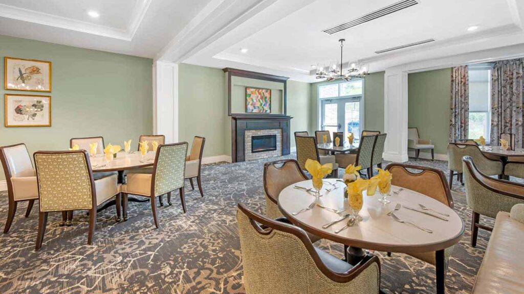 A bright, elegant dining room with round tables set with yellow napkins and silverware, upholstered chairs, patterned carpet, a fireplace, artwork, and large windows letting in natural light.