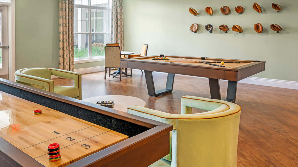 A game room with a shuffleboard table, a pool table, green upholstered chairs, a small round table, and a display of baseball gloves mounted on the pale green wall. Sunlight streams in from nearby windows.