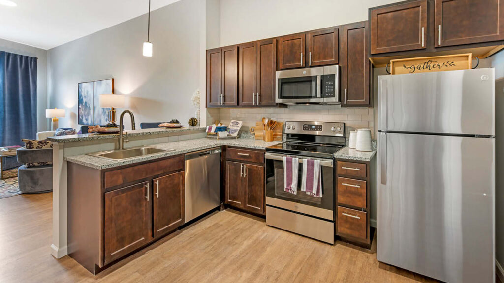 Modern kitchen with dark wood cabinets, stainless steel appliances, granite countertops, and a double sink; open layout connected to a living room with a blue curtain and contemporary decor.