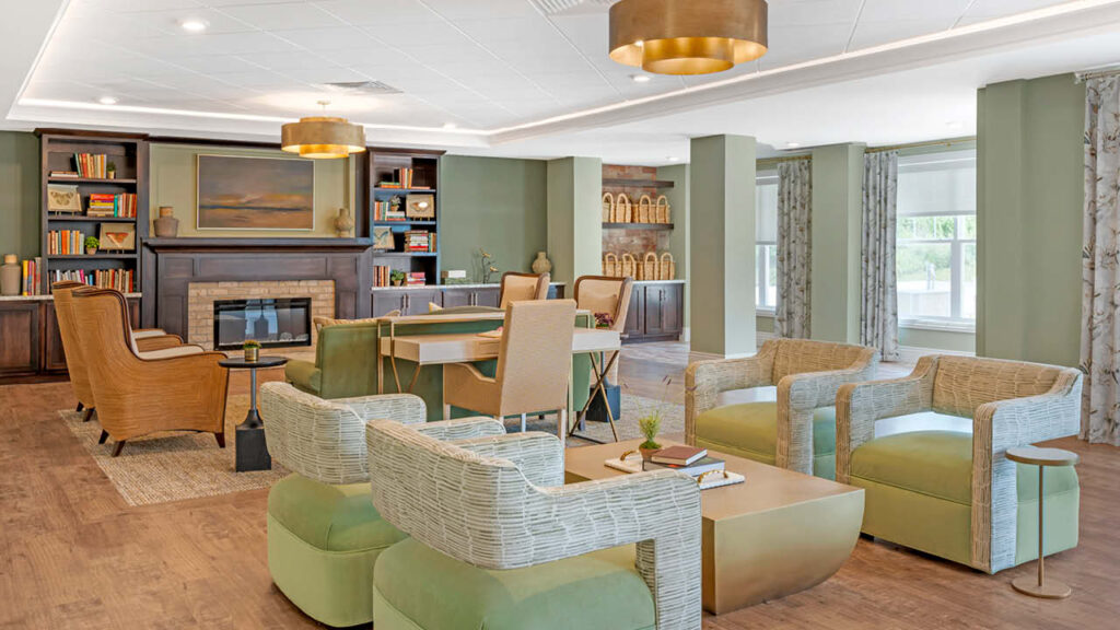 A modern lounge with green armchairs, wicker chairs, a fireplace, bookshelves, a large table, and gold ceiling lights. Large windows let in natural light, and wood floors add warmth to the spacious, inviting room.