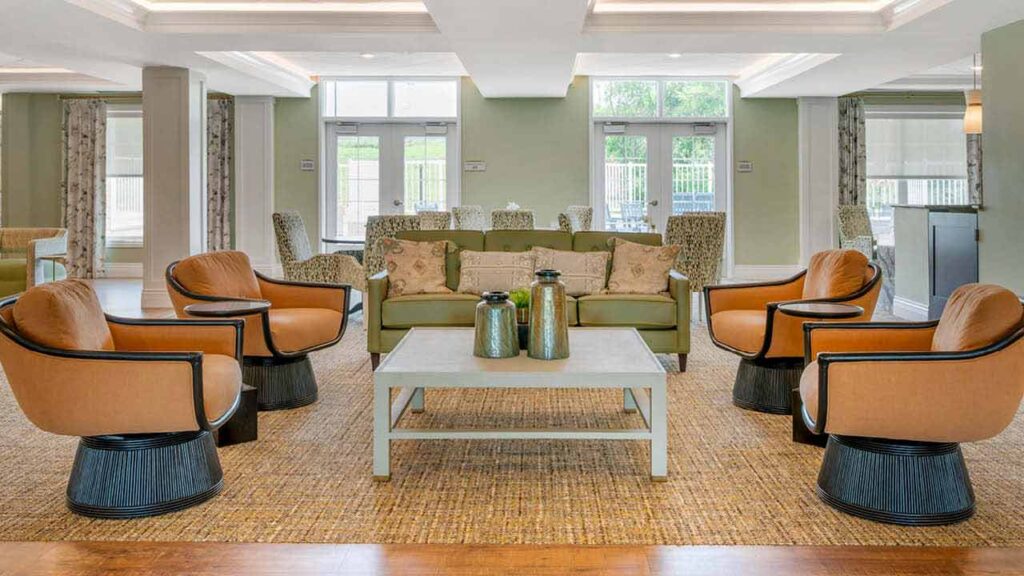 A modern lounge area with a green sofa, four tan swivel chairs, two patterned armchairs, and a white coffee table with decorative vases, all set on a woven rug in a bright room with large windows.