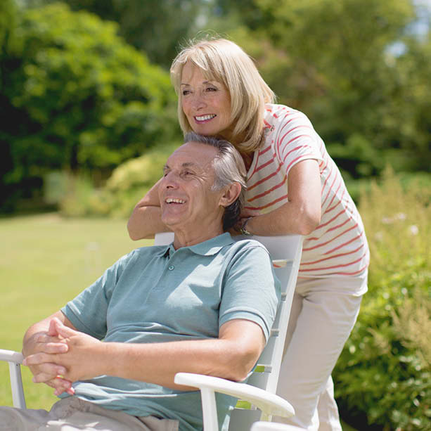 An older couple enjoys a sunny day outdoors; the man sits in a white chair smiling while the woman stands behind him with her arms around his shoulders, both looking content in a green garden setting.