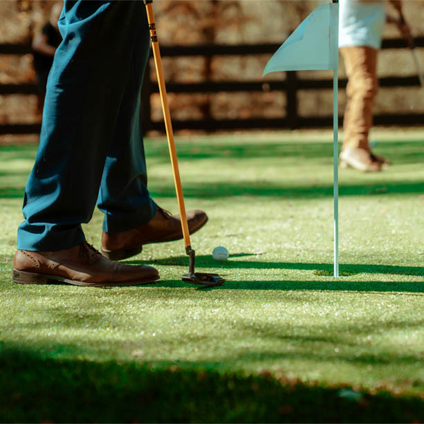 A person in dress shoes and slacks prepares to putt a golf ball near a hole on a putting green, with another person standing in the background.