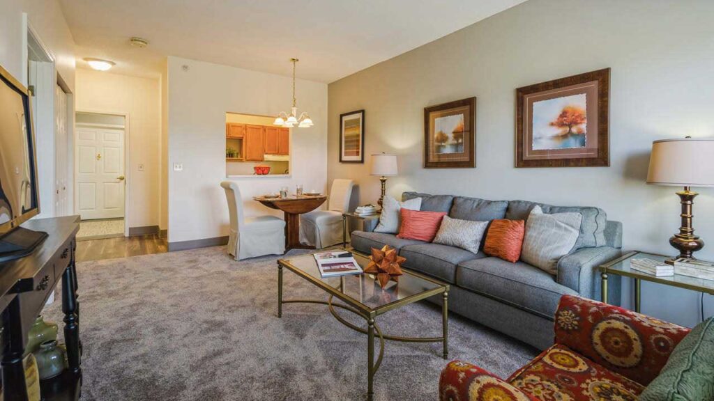 A cozy living room with a gray sofa, colorful cushions, a glass coffee table, artwork on the walls, and a dining area with a round table and two chairs near an open kitchen. Warm lighting and neutral tones create a welcoming atmosphere.