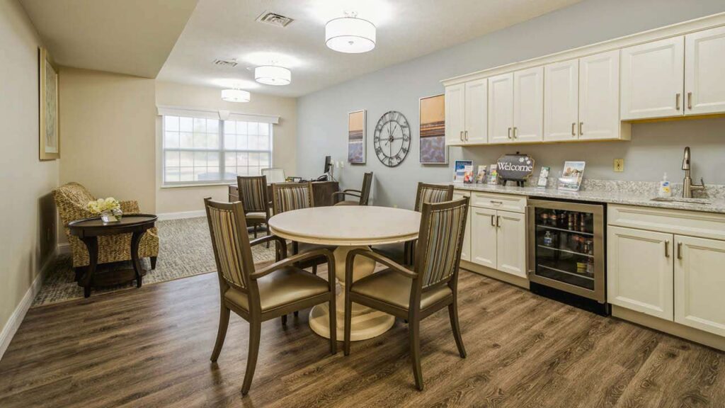 A cozy kitchen and dining area with a round table and four chairs, white cabinets, a sink, a small fridge, wall art, a large wall clock, and a sitting area with an armchair by the window.